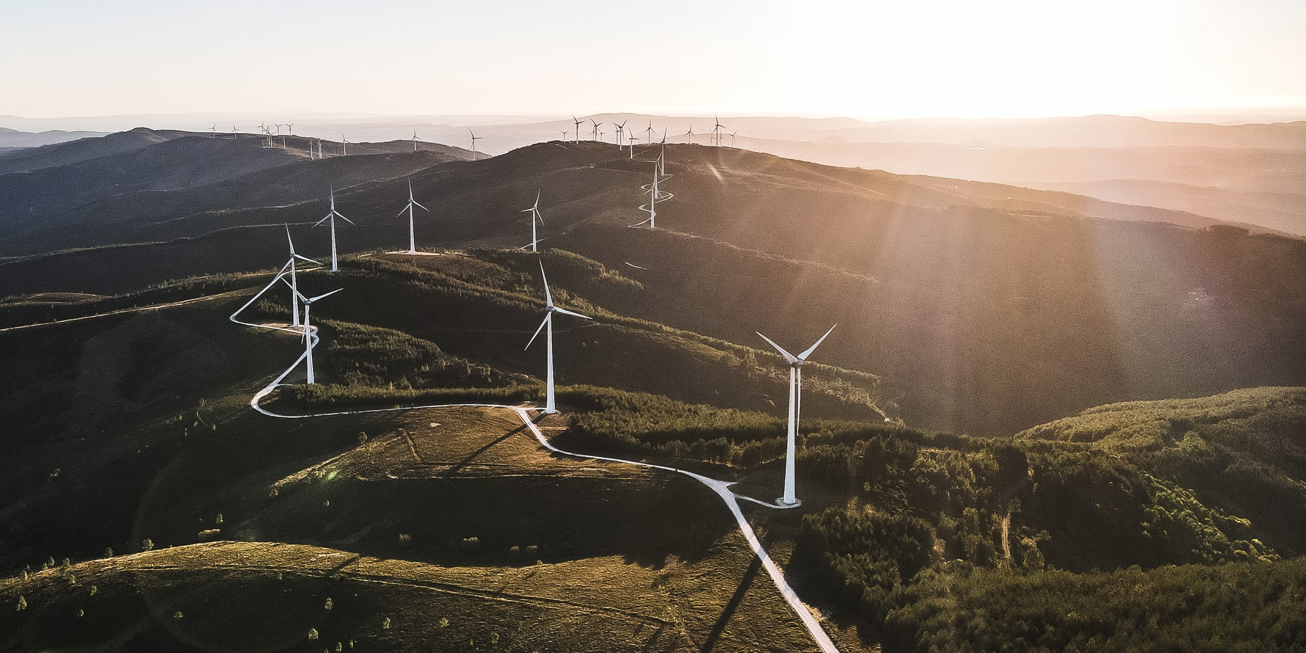 An image of windmills situated by a trail in a hilly terrain with the sun in the background.